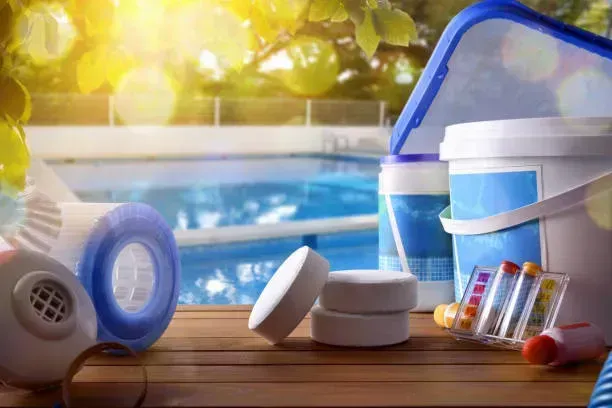 Artículos de natación y protector solar sobre una mesa de madera junto a una piscina azul.