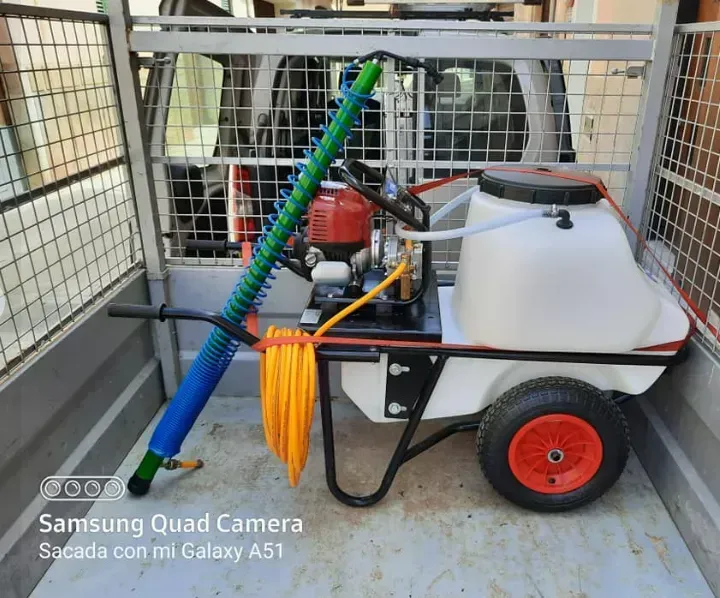 Unidad pulverizadora portátil con manguera, rueda roja y tanque blanco en la caja de una camioneta.