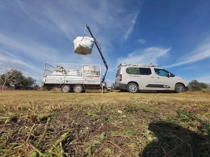 Furgoneta blanca remolcando un pequeño remolque que levanta una paca de heno en un campo de hierba bajo un cielo azul.