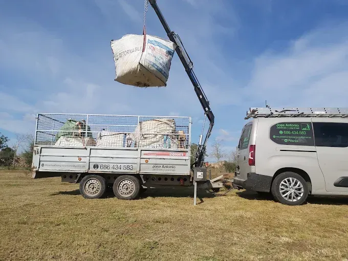 Una grúa carga sacos blancos en un camión de plataforma junto a una furgoneta blanca en un campo de hierba.