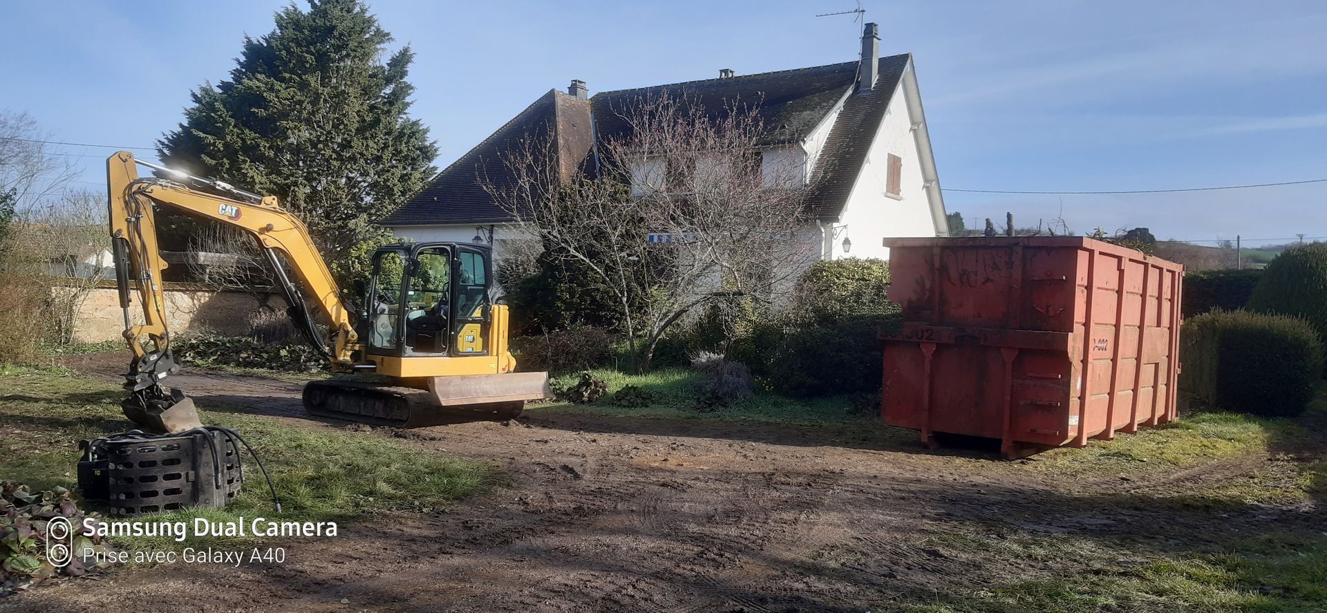 Pelleteuse jaune sur un terrain avec une maison blanche et un conteneur
