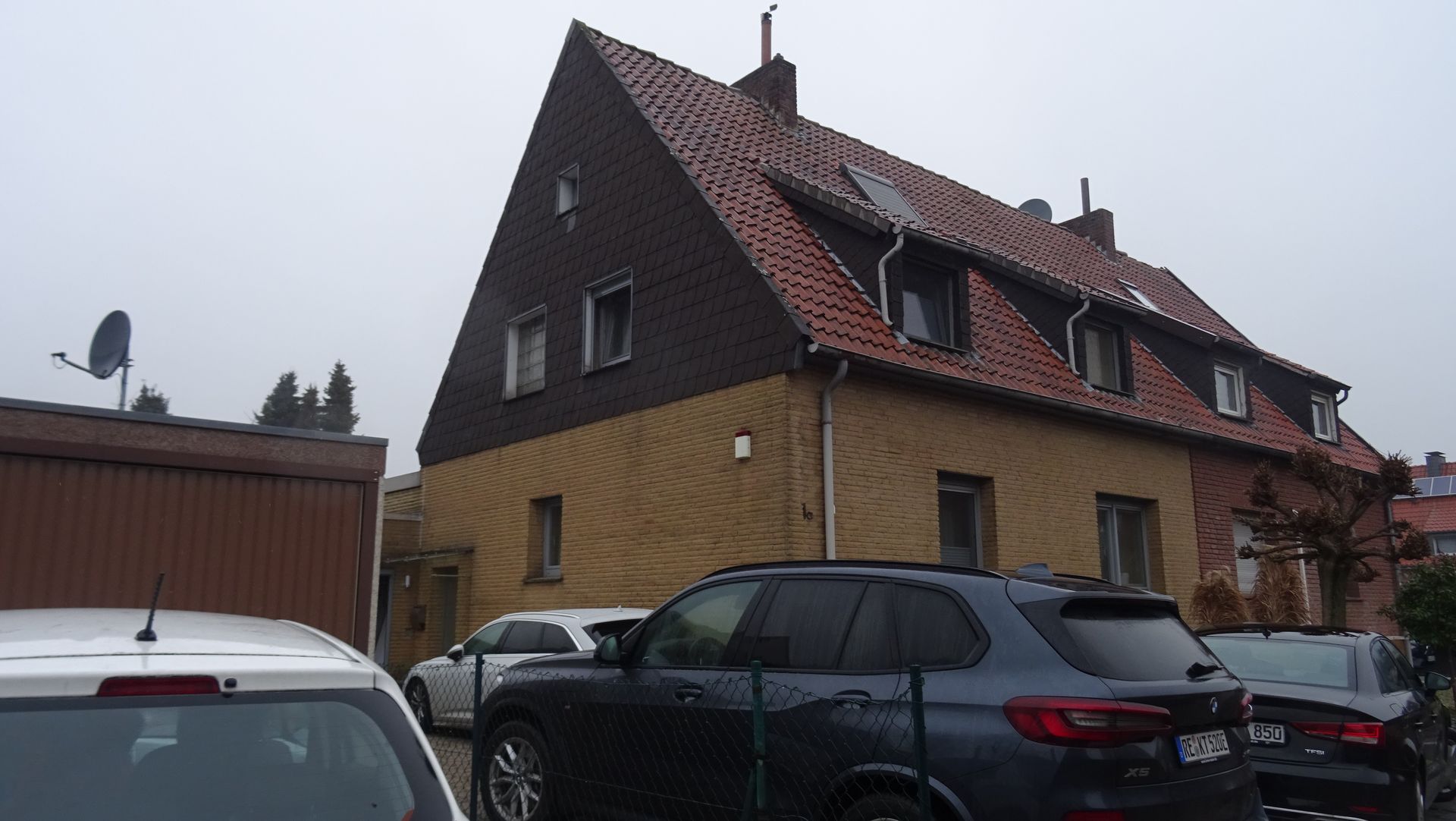 Geparkte Autos neben einem Backsteinhaus mit steilem, rotem Ziegeldach an einem bewölkten Tag