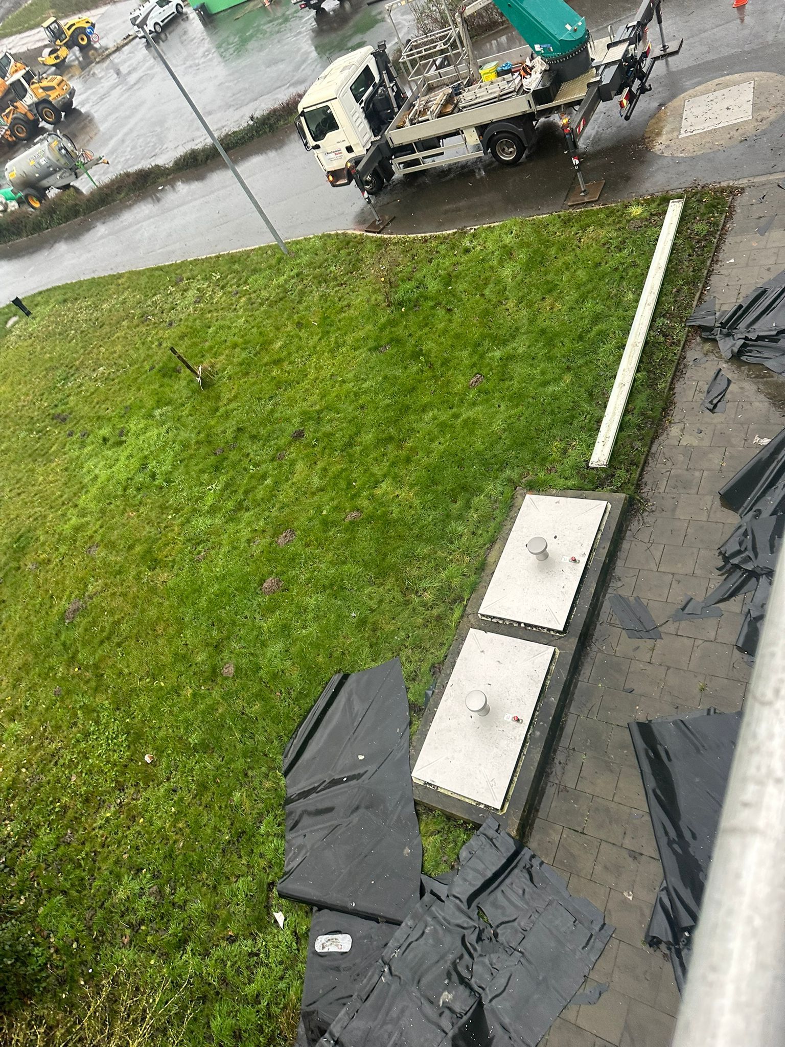 Eine regnerische Straße neben einem grasbewachsenen Hang, darunter Baumaschinen und Verteilerkästen.