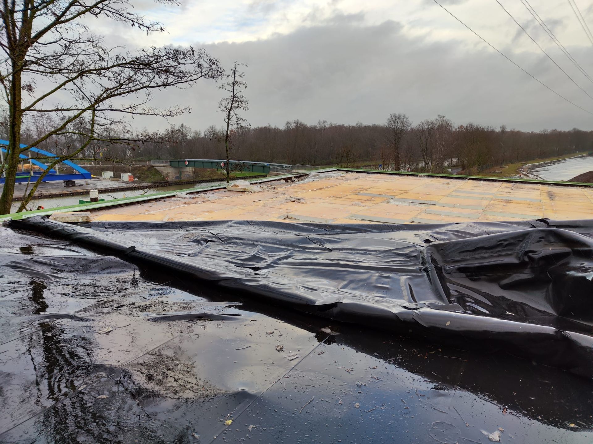 Überschwemmter Straßenrand mit einer zerrissenen schwarzen Plane oder Auskleidung auf schlammigem Wasser unter bewölktem Himmel.