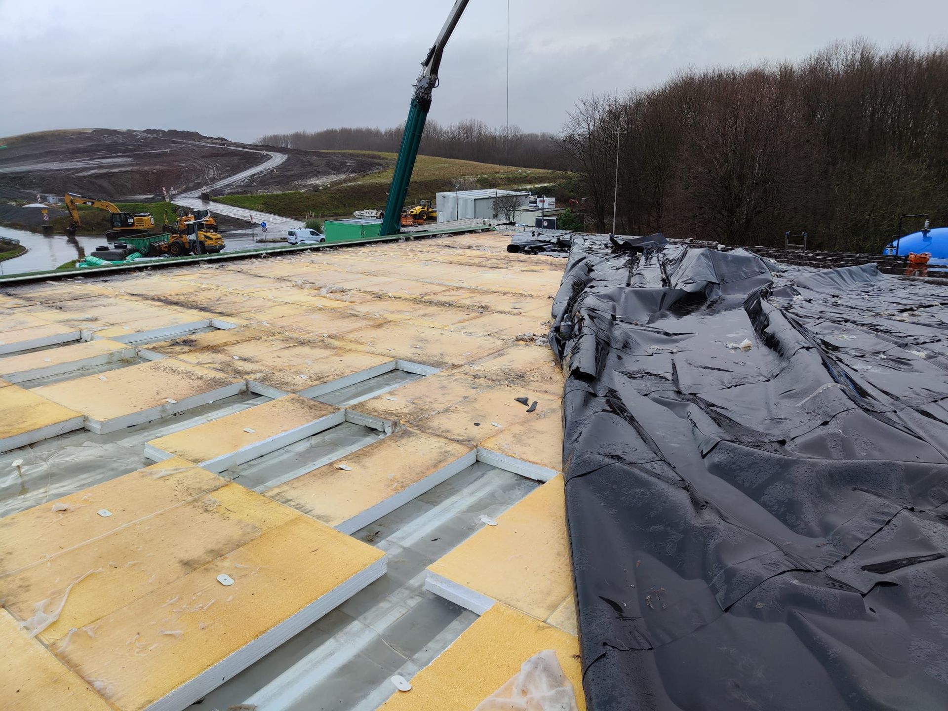 Baustelle mit gelben Dämmplatten und einer großen schwarzen Plane auf einem Dach, im Hintergrund ein Kran.