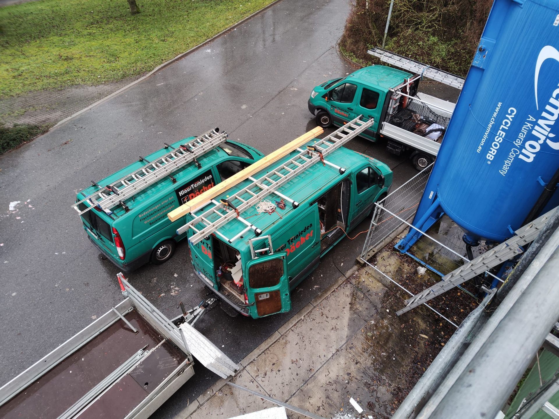 Umgestürzte türkisfarbene Lieferwagen blockieren eine nasse Straße neben einem blauen Industriesilo.