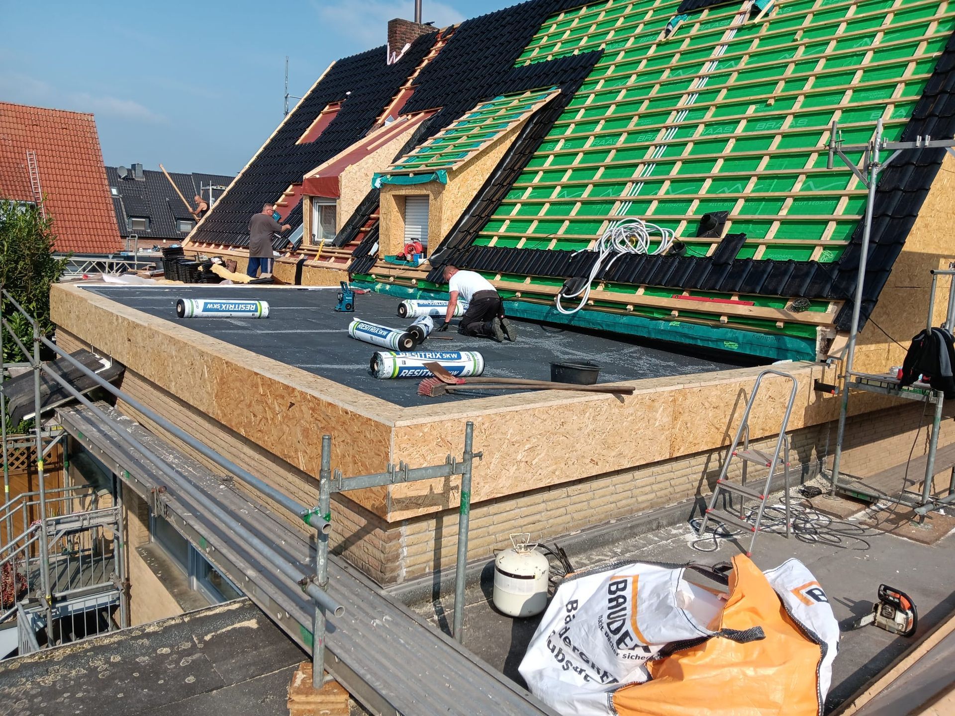Auf einem eingerüsteten Haus ist ein Dach im Bau, auf dem Arbeiter, grüne Ziegel und Baumaterialien zu sehen sind.
