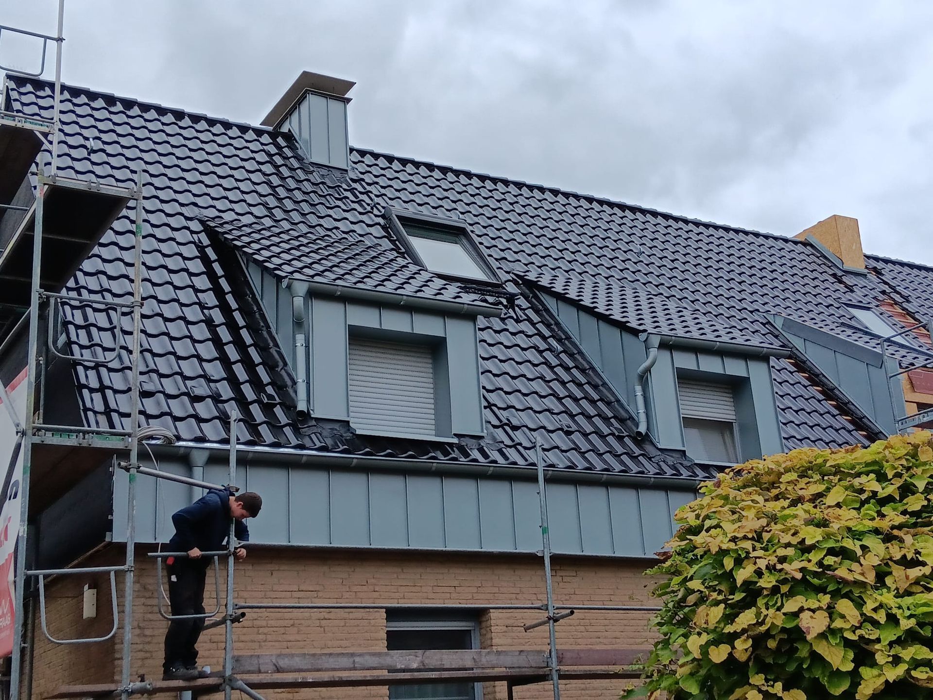 Ein Arbeiter repariert auf einem Gerüst ein graues Ziegeldach mit Dachgauben.