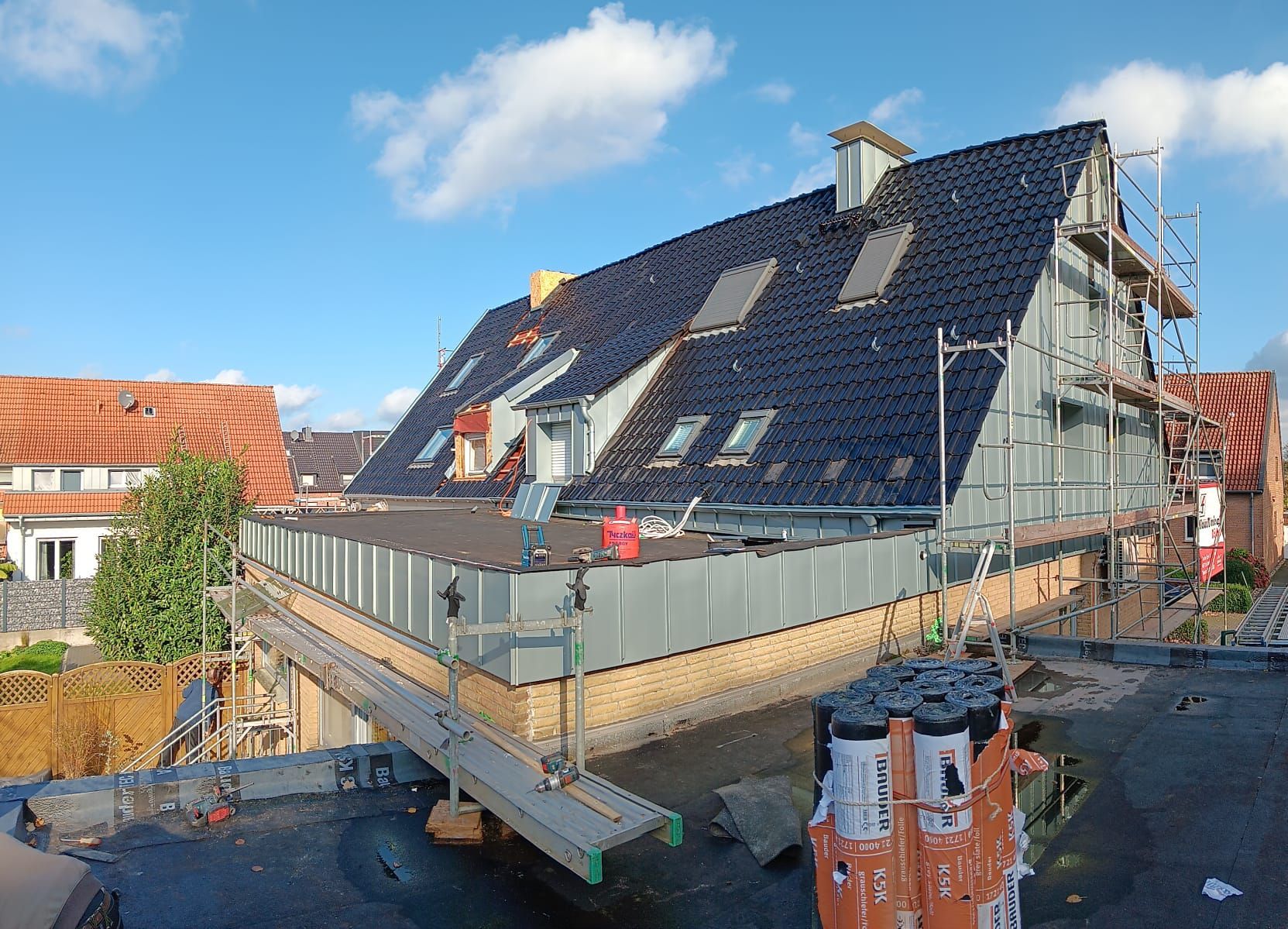 Das Dach wird gerade umgebaut, ein Gerüst steht darauf, und unter blauem Himmel steht ein schräges, dunkel geziegeltes Haus.