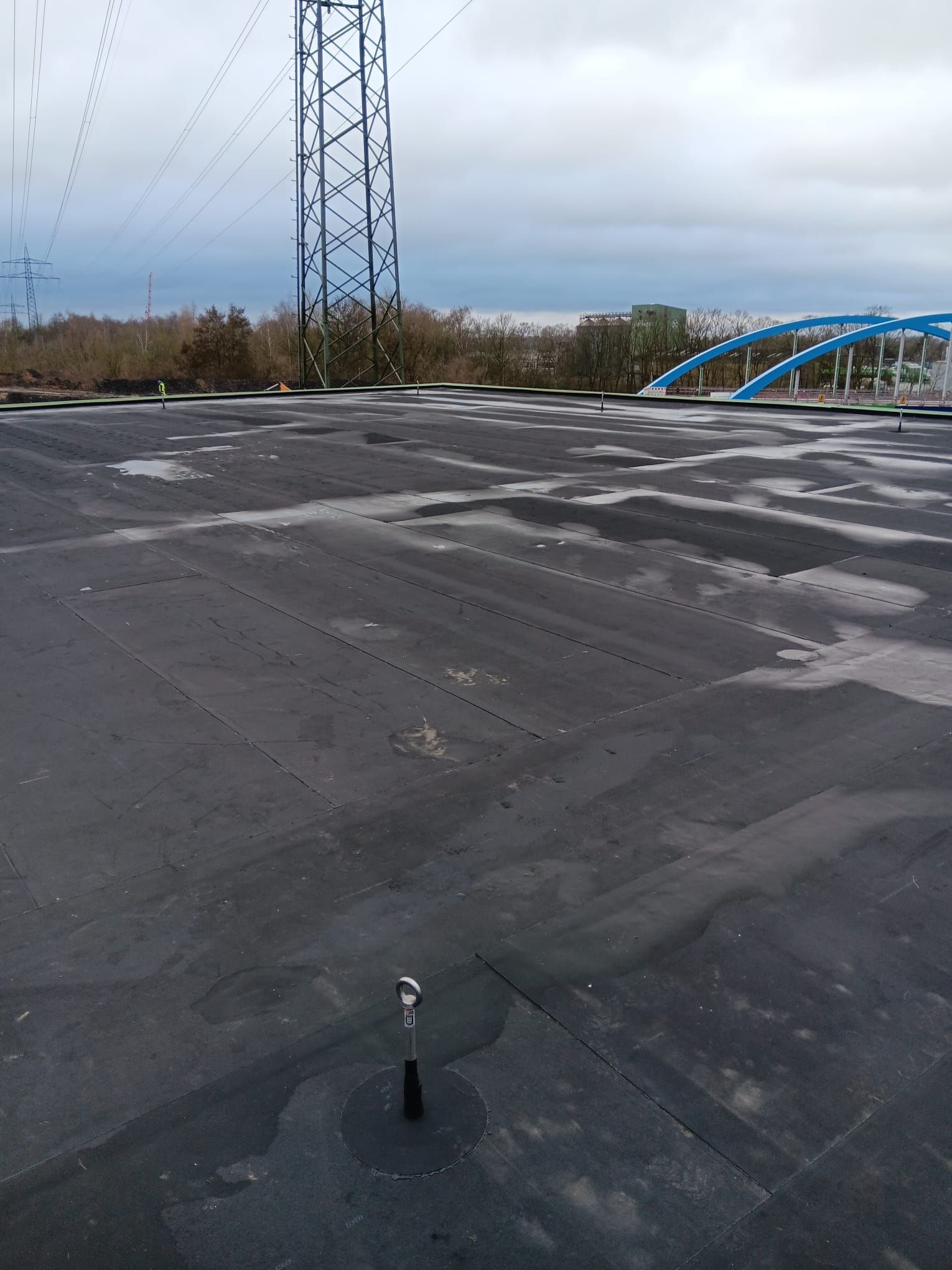 Ein Dach mit dunklem Asphalt, ein Metallturm und in der Ferne eine blaue Bogenbrücke unter bewölktem Himmel