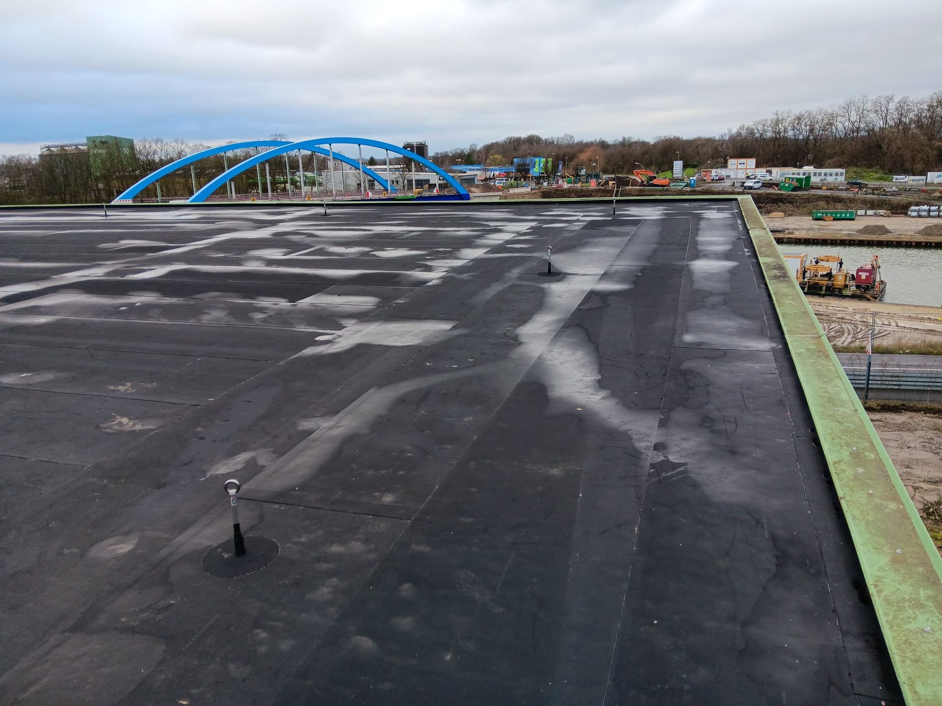 Dachterrasse mit schwarzer wasserdichter Membran und in der Ferne eine blaue Bogenbrücke unter bewölktem Himmel