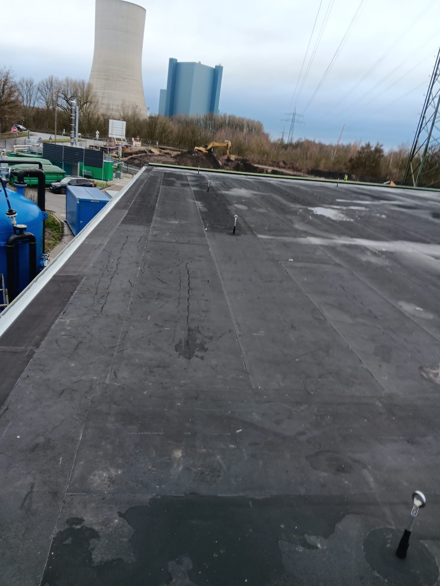 Flachdach mit dunkler, wasserdichter Membran, Lüftungsöffnungen und in der Ferne Industriegebäude unter bewölktem Himmel