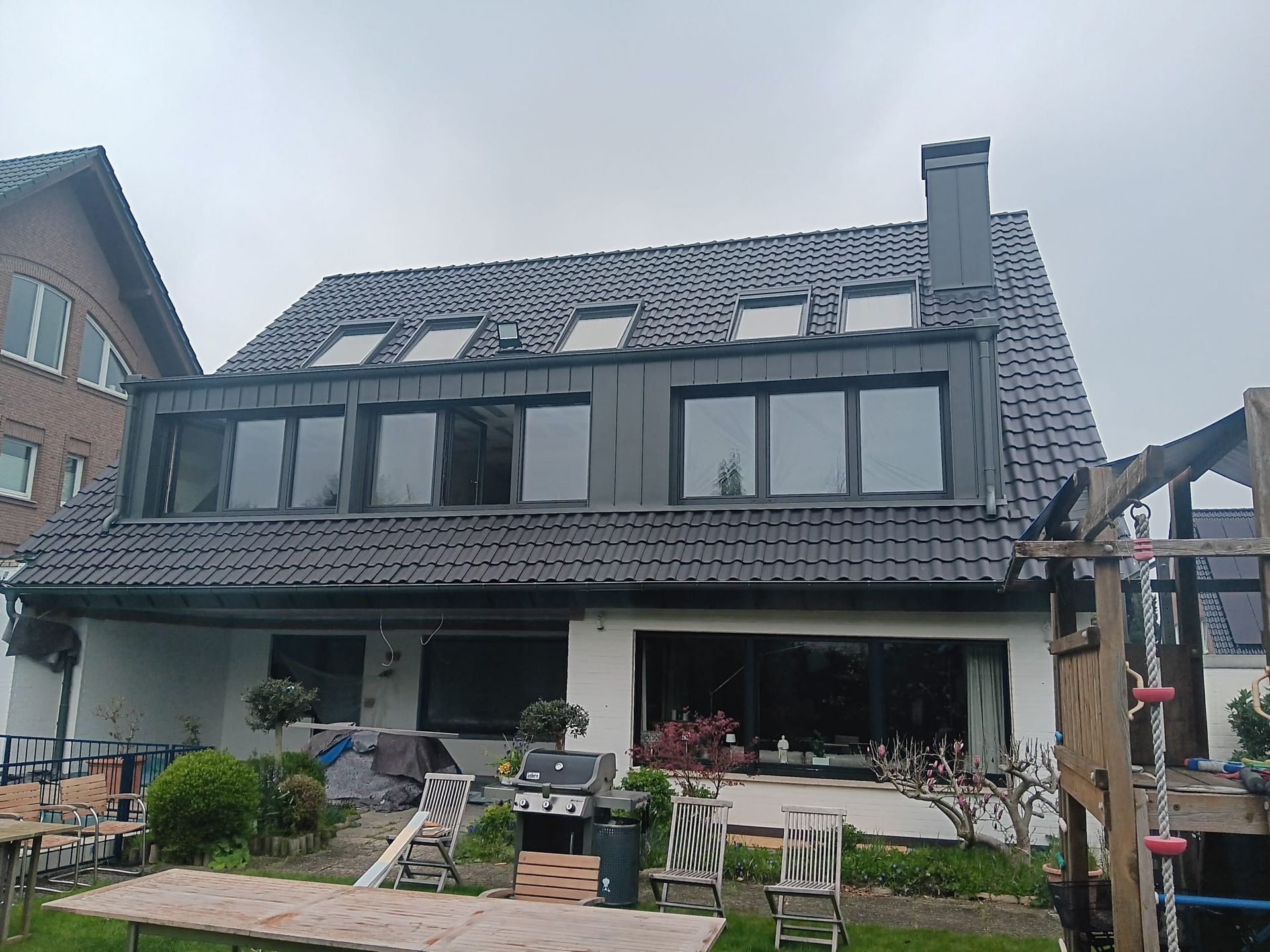Modernes zweistöckiges Haus mit grauem Ziegeldach, Dachgauben und einem kleinen Vorgarten mit Stühlen