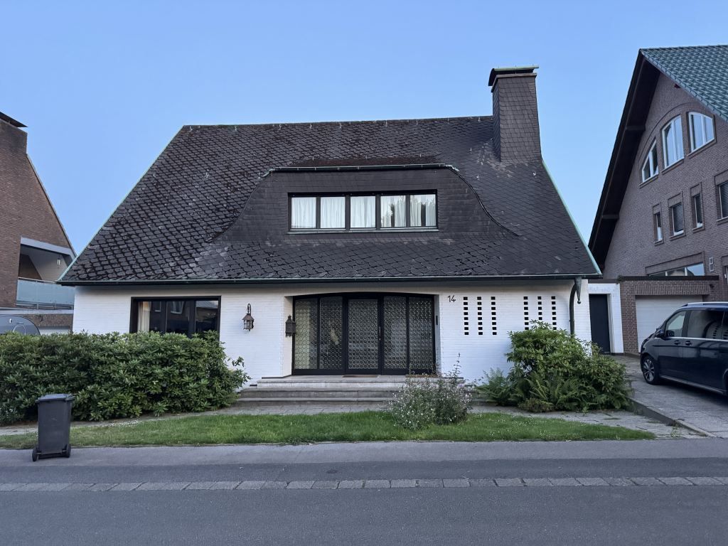 Haus mit dunklem Dach und weißer Fassade in einer Wohnstraße, flankiert von Nachbarhäusern.
