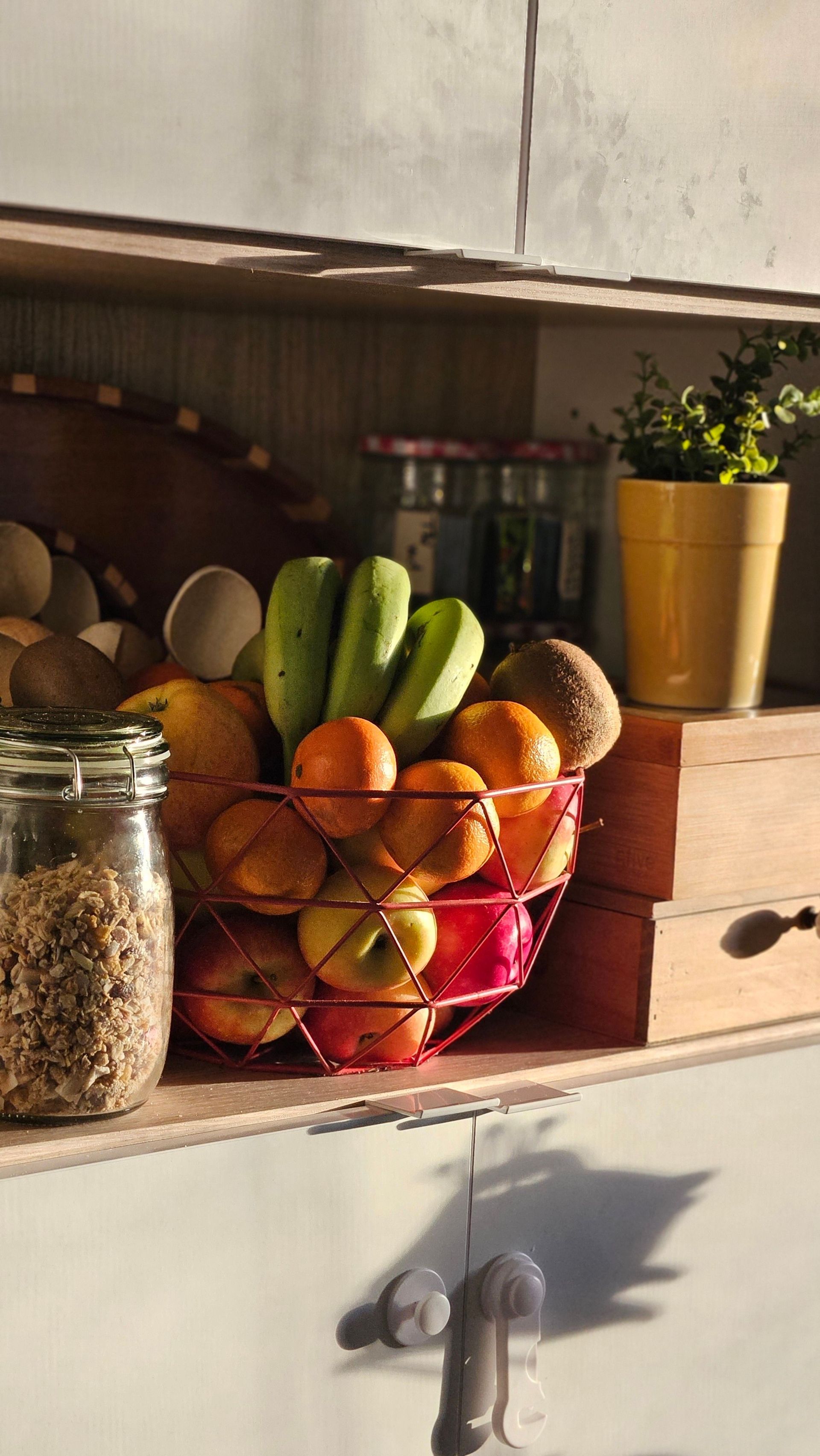 Étagère de cuisine avec un panier en fil de fer rempli de fruits, un bocal de céréales et une plante en pot dans un pot jaune.