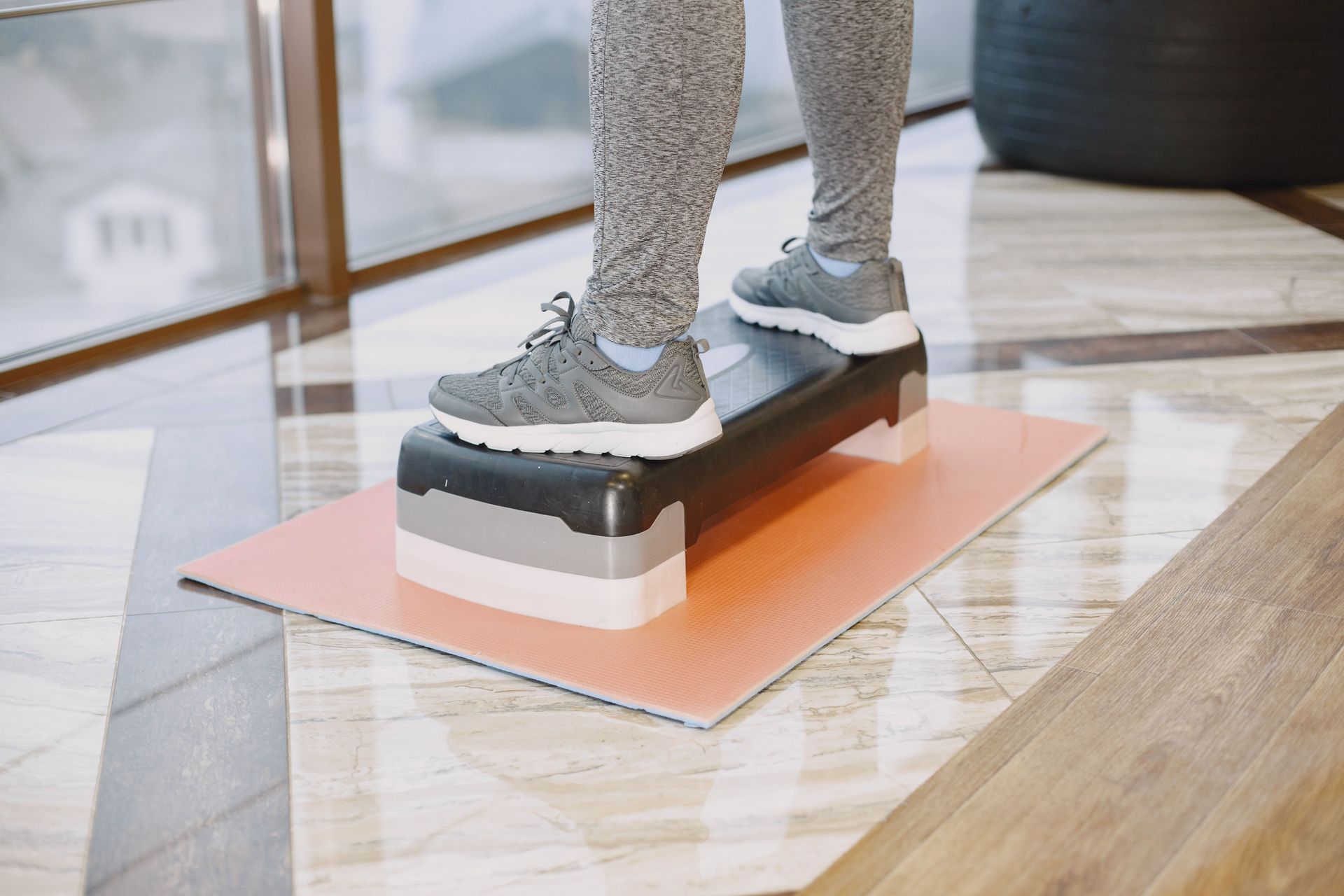 Une personne se tient en équilibre sur une planche dans une pièce intérieure lumineuse, vêtue d'un legging gris et de baskets.