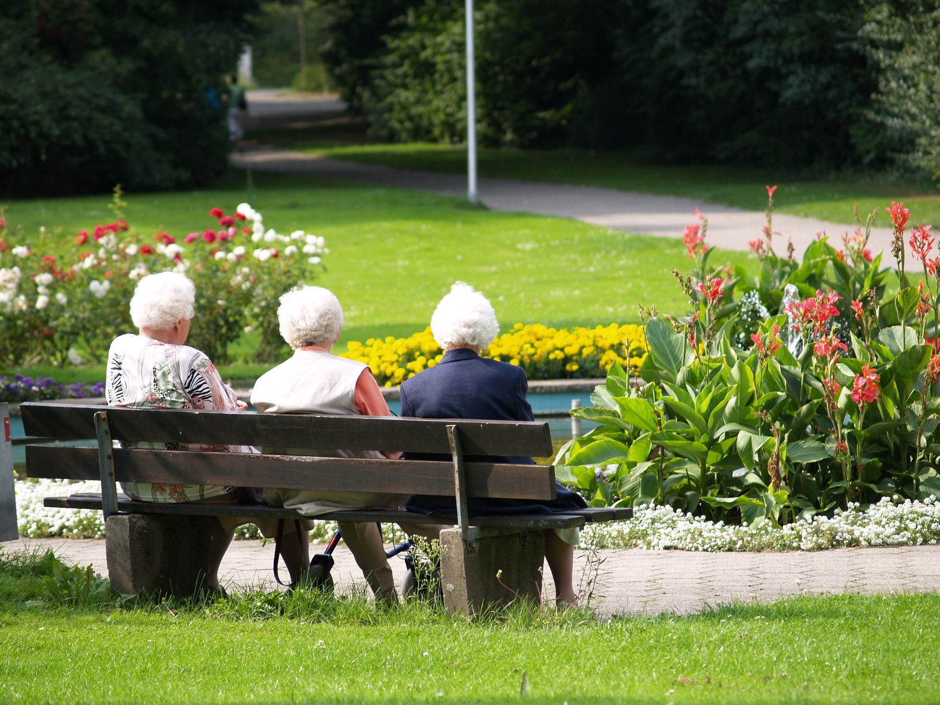 Trois personnes âgées sont assises sur un banc, face à un jardin fleuri dans un parc.