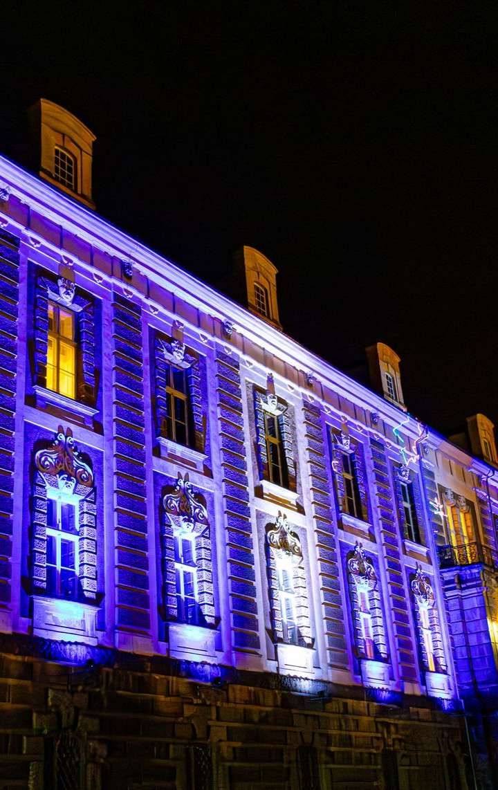 La façade du bâtiment est illuminée en violet et blanc la nuit.