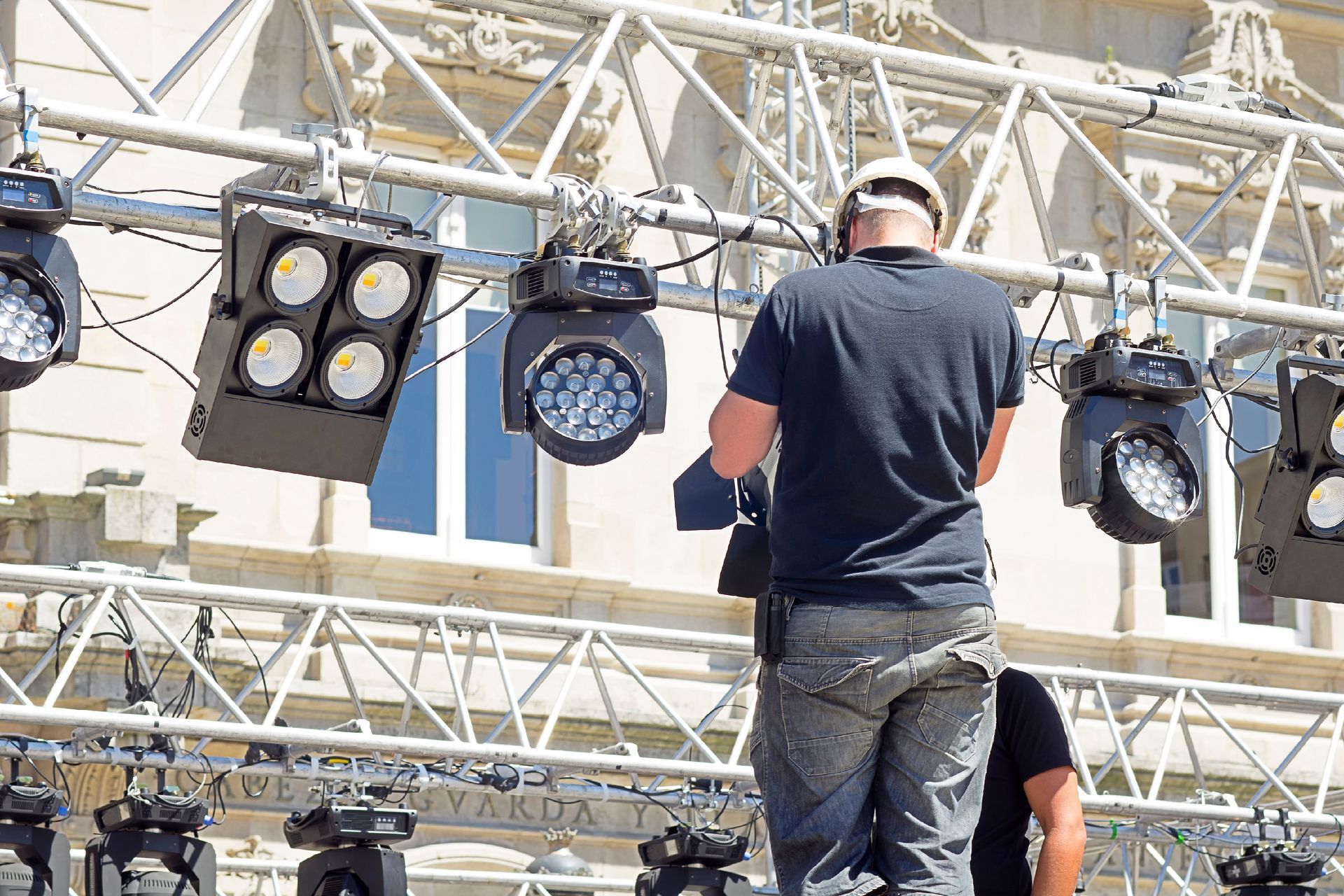 Une personne portant un casque de chantier travaille sur des projecteurs de scène, en extérieur, par une journée ensoleillée.