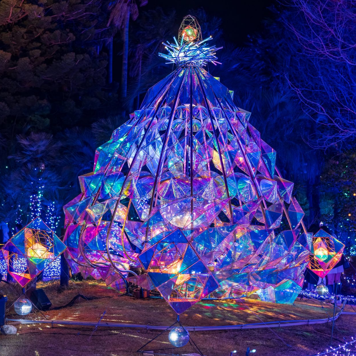 Installation artistique géante, illuminée et prismatique, en forme d'arbre et composée de panneaux irisés. Installée en extérieur, la nuit.
