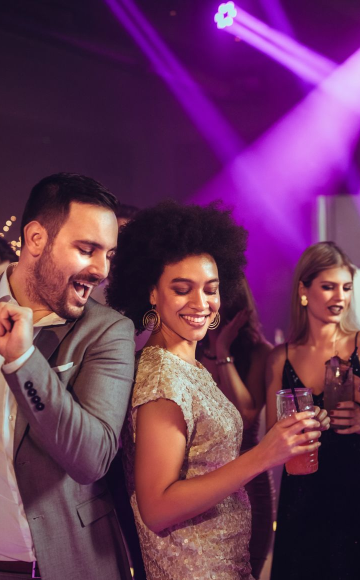 Des personnes dansent lors d'une fête sous des lumières de scène violettes. Une femme en robe dorée sourit, un verre à la main, à côté d'un homme.