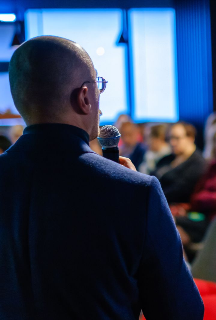 Un homme parle au micro, face au public. Éclairage bleu et rouge dans la salle.
