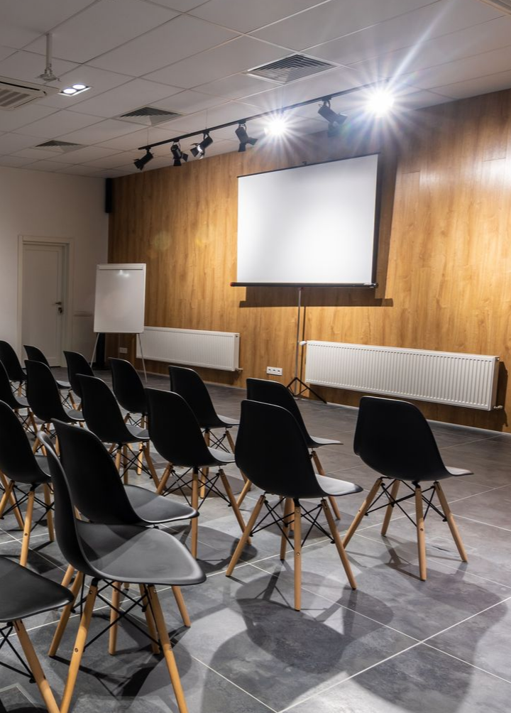 Salle de conférence vide avec des chaises noires face à un écran de projection vierge, un mur en bois et un éclairage zénithal.