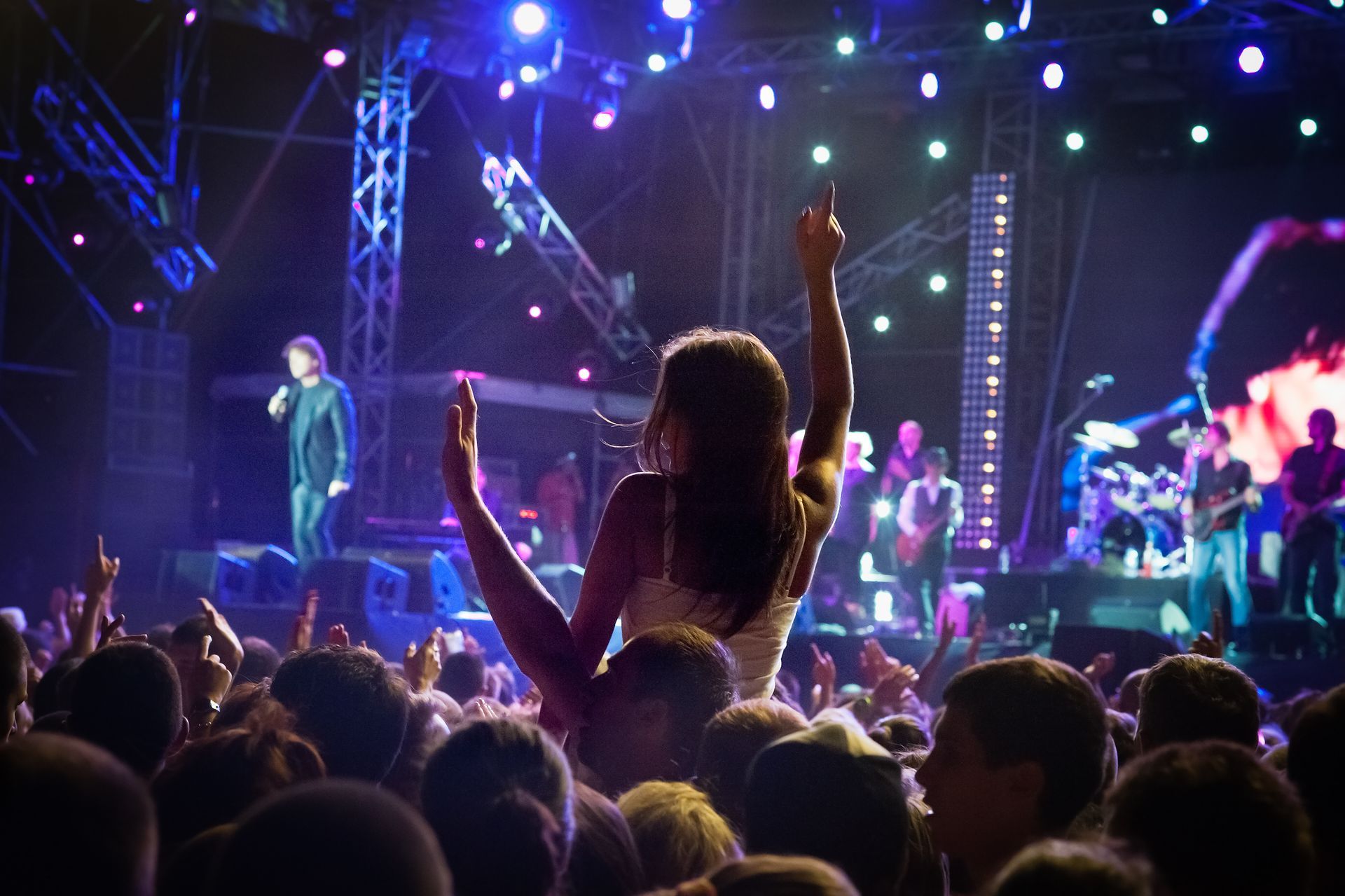 Une femme portée sur les épaules lors d'un concert, les bras levés. Scène avec le groupe en arrière-plan, lumières vives.