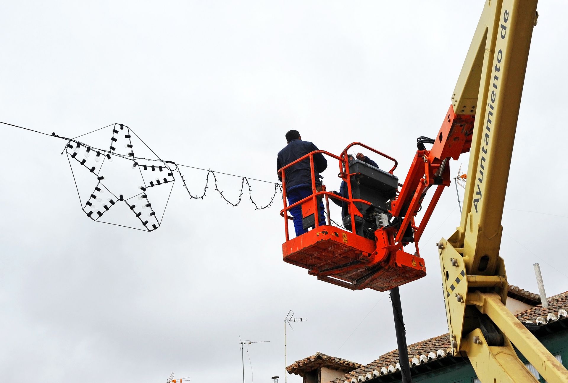 Un ouvrier dans une nacelle installe des décorations d'étoiles de Noël dans une rue. Ciel nuageux.