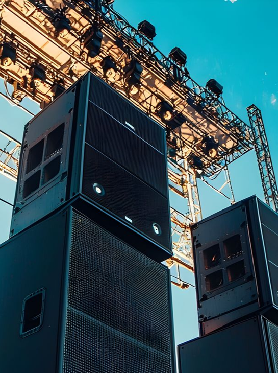 Des enceintes de concert noires empilées, du matériel de scène illuminé sur fond de ciel bleu.