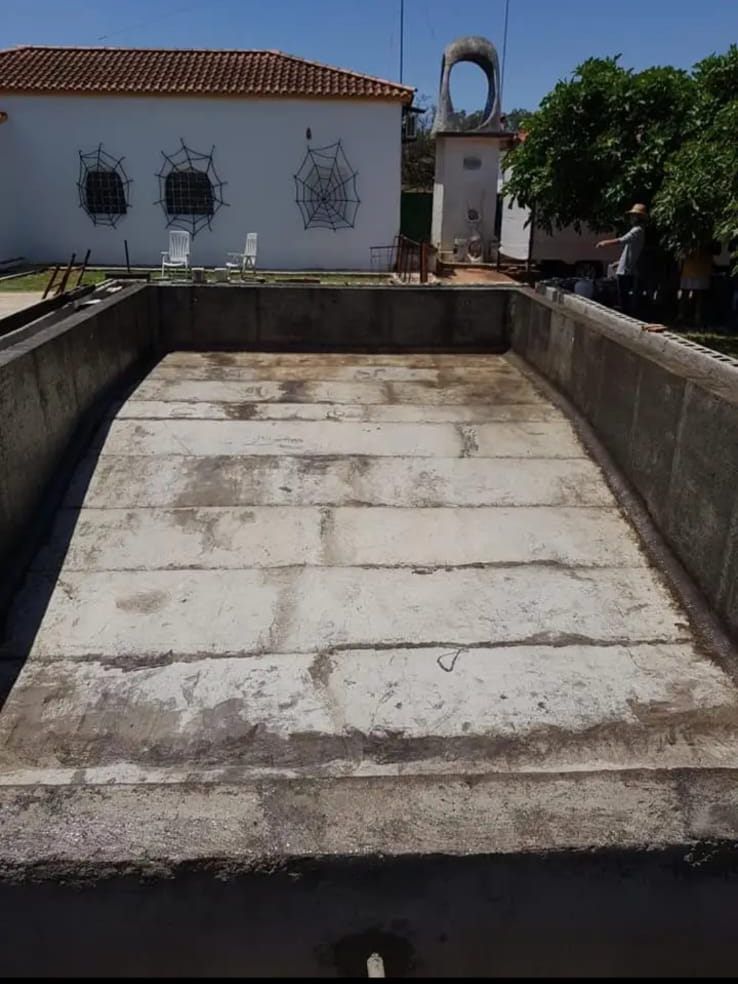 Piscina de hormigón vacía con baldosas rectangulares, frente a un edificio blanco con una torre, exterior soleado.