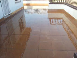 Un patio exterior húmedo y embaldosado refleja la luz. Se ven azulejos marrones, barandilla blanca y edificios.