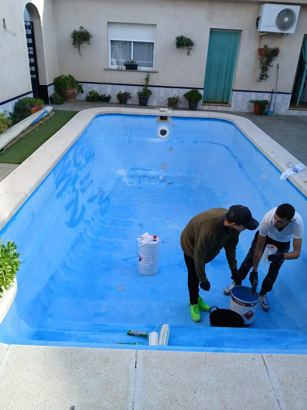Dos personas pintan una piscina azul vaciada. Están en un patio.