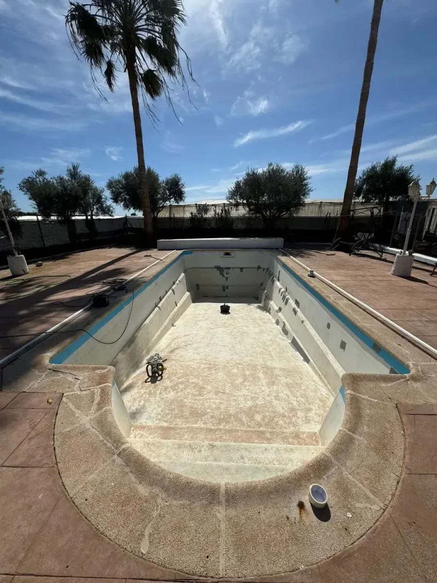 Piscina vacía y agrietada, rodeada de hormigón. Palmeras y cielo azul visibles al fondo.