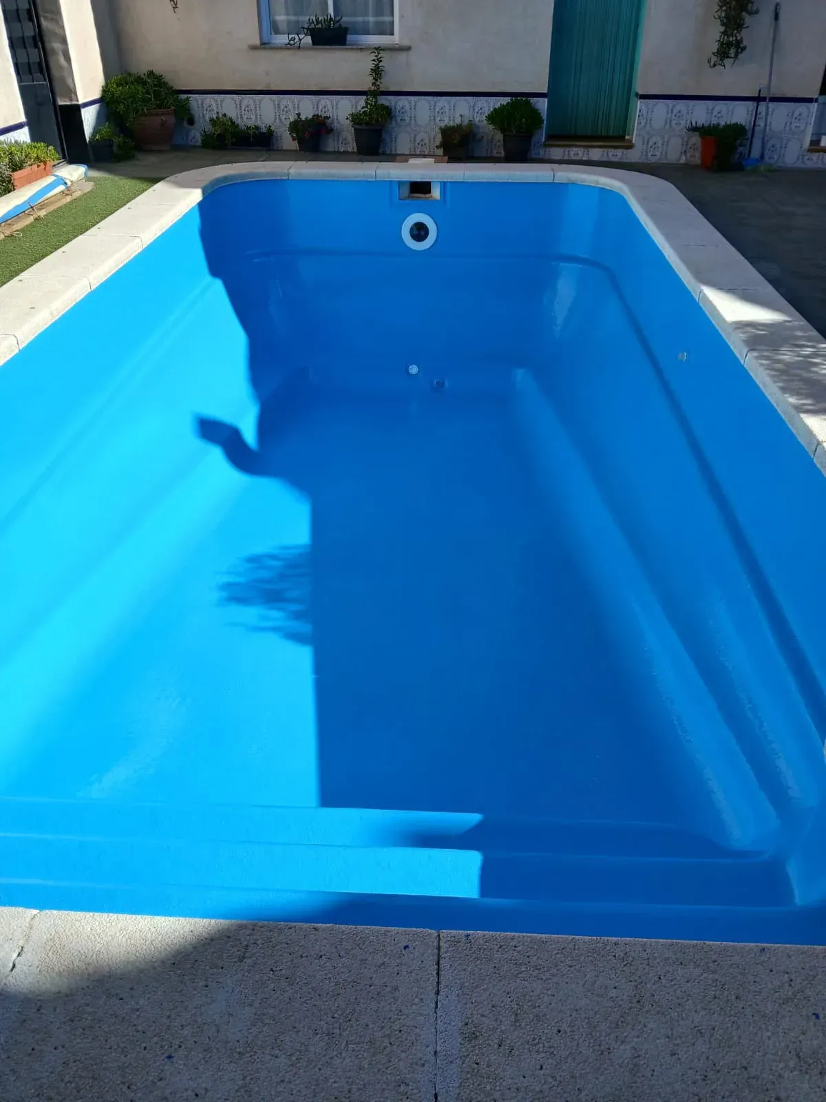 Piscina rectangular azul vacía, escalones, cubierta de hormigón claro y plantas verdes.