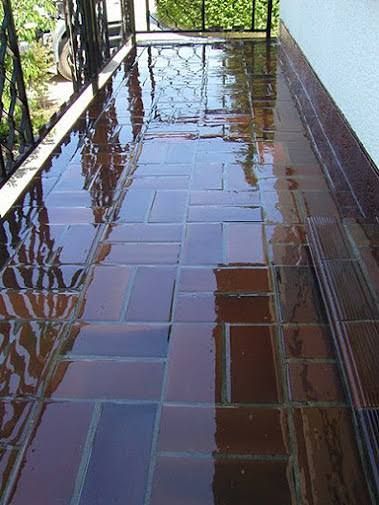 Porche húmedo, de baldosas marrones, que refleja el entorno, barandilla de hierro a la izquierda, pared blanca a la derecha.