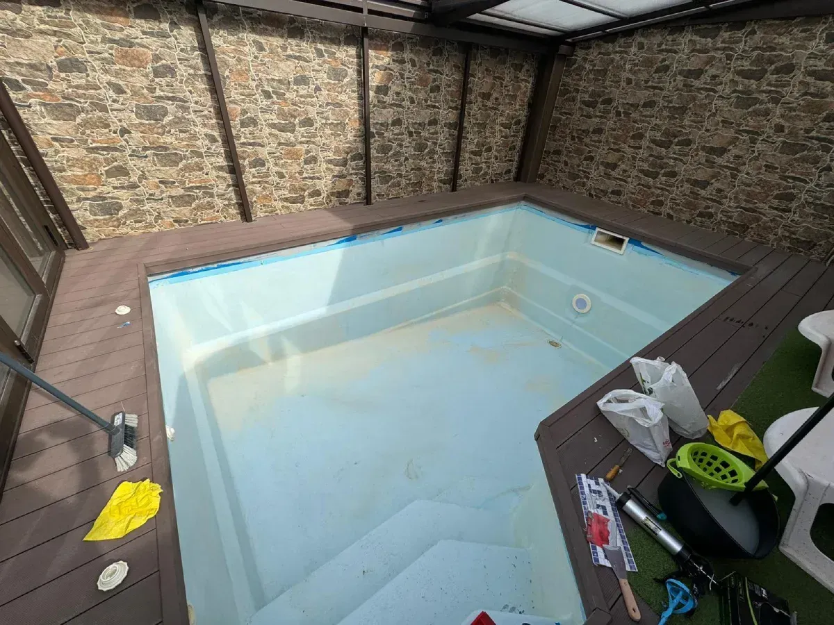 Pequeña piscina rectangular vacía con tarima marrón, contra una pared de piedra texturizada, bajo un techo translúcido.