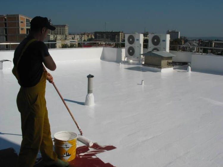 Persona aplicando pintura blanca a un tejado plano con rodillo y balde; entorno urbano.