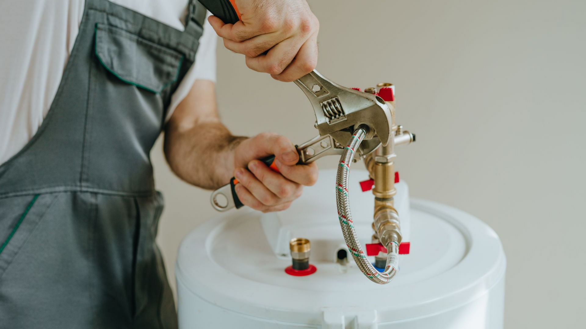 Un plombier en salopette grise utilise une clé pour raccorder un tuyau flexible à un chauffe-eau blanc.