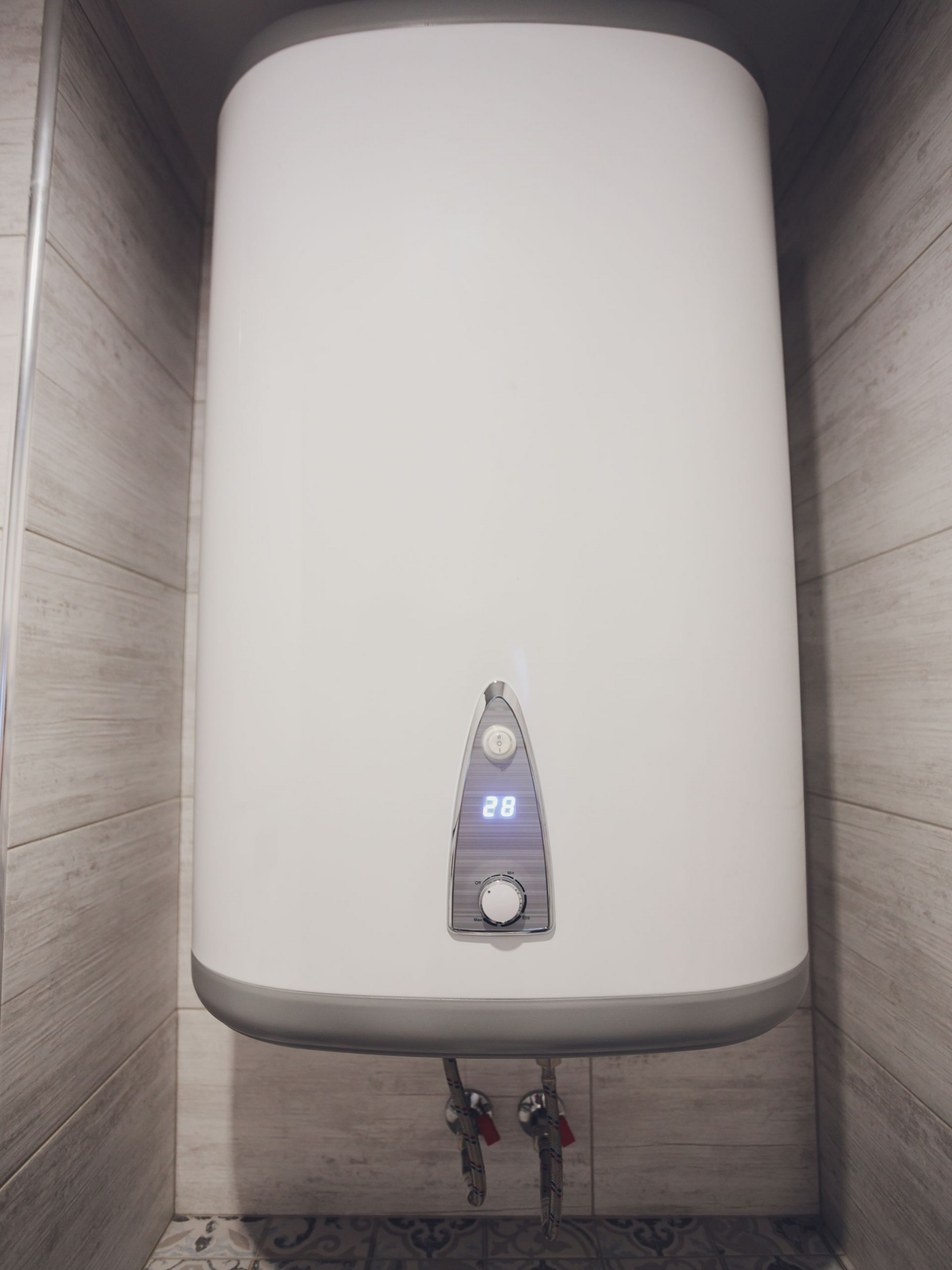 Chauffe-eau blanc fixé sur un mur carrelé, avec panneau de commande et tuyaux d'eau.