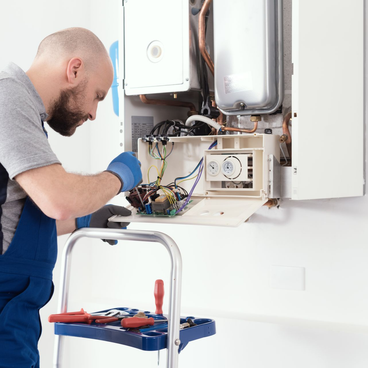 Un plombier en salopette bleue répare une chaudière blanche à l'aide d'outils.
