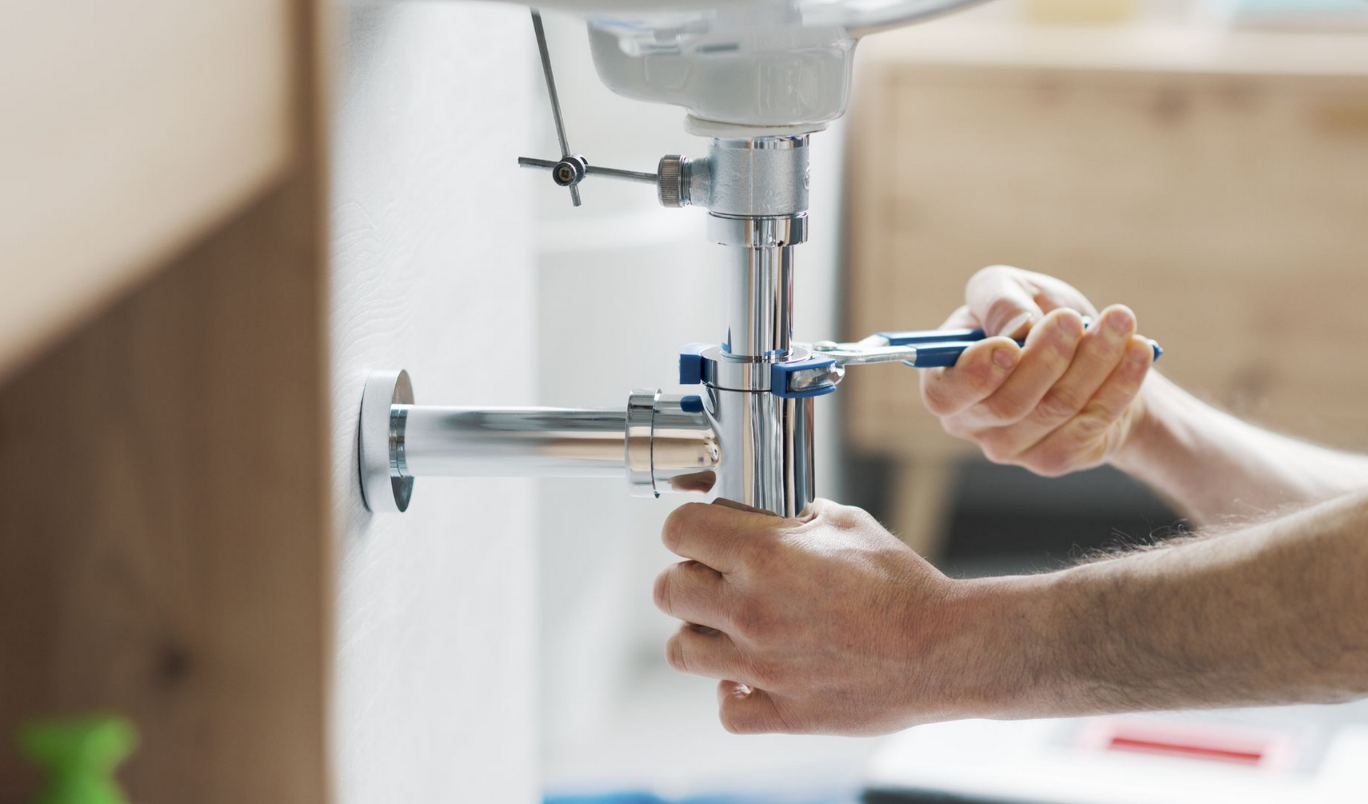 Des mains utilisent une clé pour resserrer la tuyauterie sous un lavabo blanc dans une salle de bains.