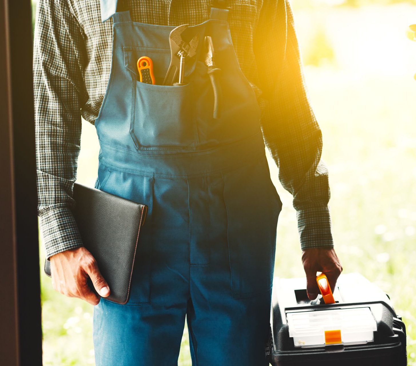 Une personne en salopette bleue sur le palier, tenant une boîte à outils et un dossier.