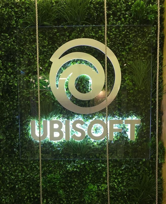 Le logo Ubisoft en blanc, rétroéclairé sur un panneau transparent fixé contre un mur végétal luxuriant de type jardin vertical.