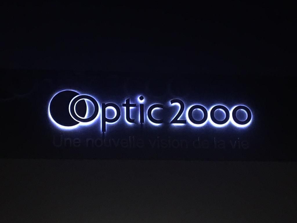 Enseigne lumineuse portant l'inscription Optic 2000 avec le slogan Une nouvelle vision de la vie sur fond sombre.