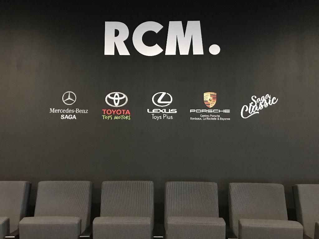 Mur portant l'inscription RCM et cinq logos automobiles au-dessus d'une rangée de sièges gris dans une salle d'attente moderne et sombre.