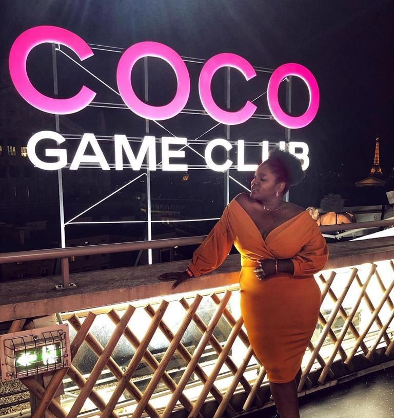Une personne vêtue d'une robe orange à épaules dénudées est appuyée contre une rambarde devant une enseigne rose vif COCO GAME CLUB.