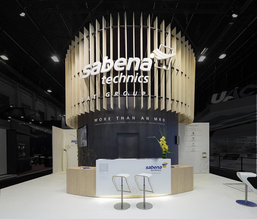 Un stand moderne pour Sabena Technics lors d'un salon professionnel, avec une grande structure cylindrique à lattes de bois et un comptoir d'accueil.