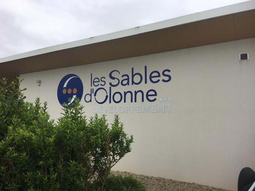 L'extérieur d'un bâtiment blanc affichant le logo bleu et le texte les Sables-d'Olonne avec des buissons verts au premier plan.
