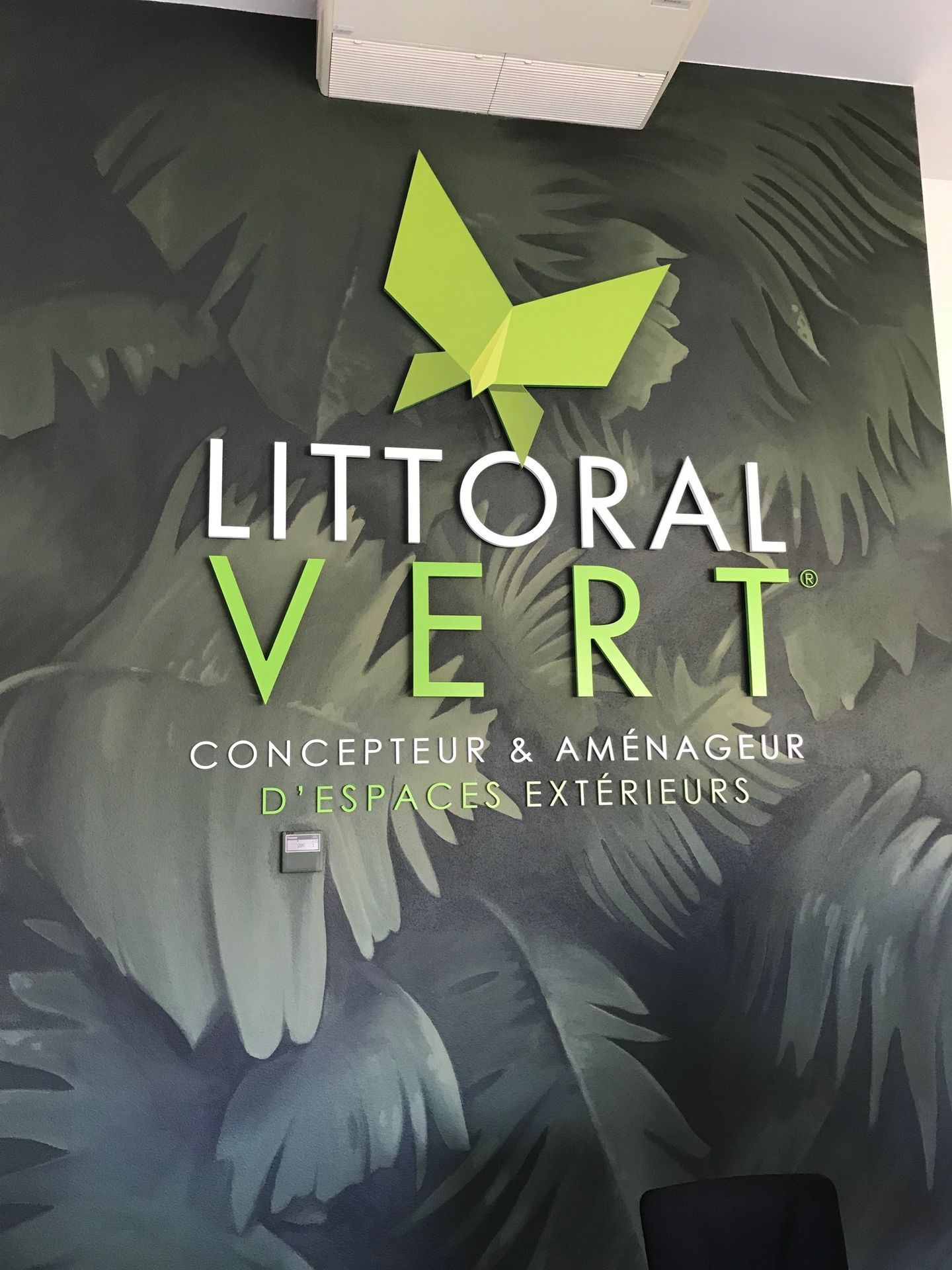 Une pancarte murale indique LITTORAL VERT sous un logo papillon vert imitant du papier, sur un papier peint à motifs de feuilles tropicales vert foncé.